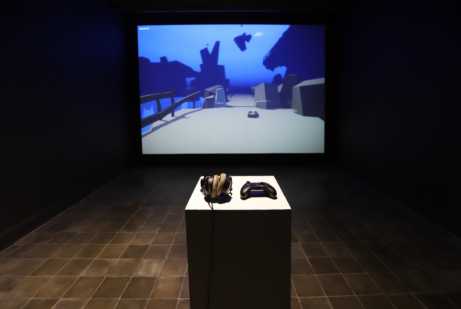 Trapped installed at ARTJOG — headphones and controller on a plinth, in-game render projected on the wall behind.